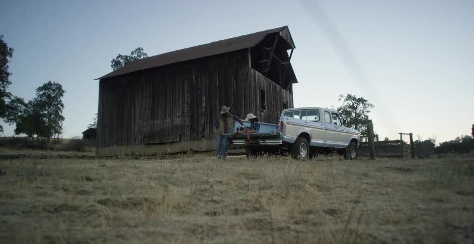 A white pick up truck is parked near a barn and a little girl is sitting in the back with a dog as a man stands next to them