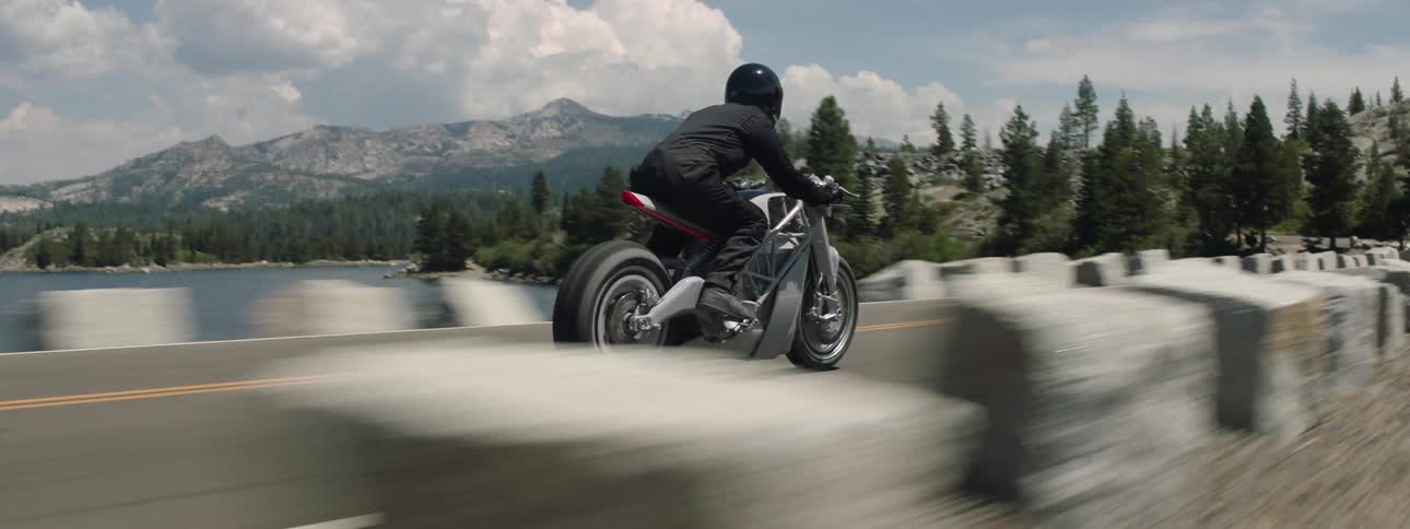 Motorcyclist rides along lakeside highway passing parked cars and curving toward forest