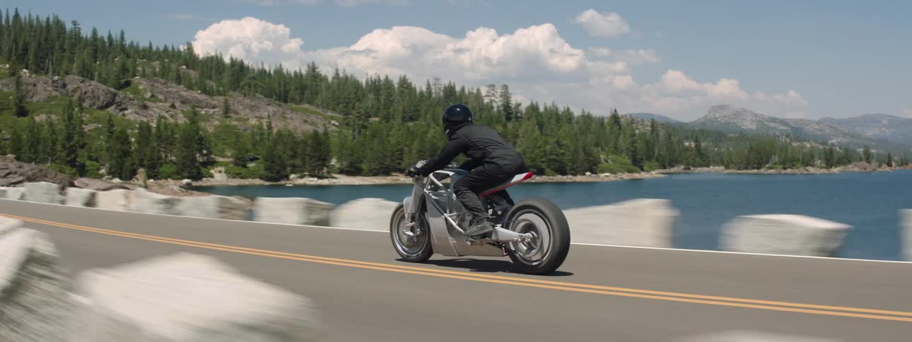 A motorcycle travels across a bridge toward a forested mountain as the rider is wearing a black jacket and helmet