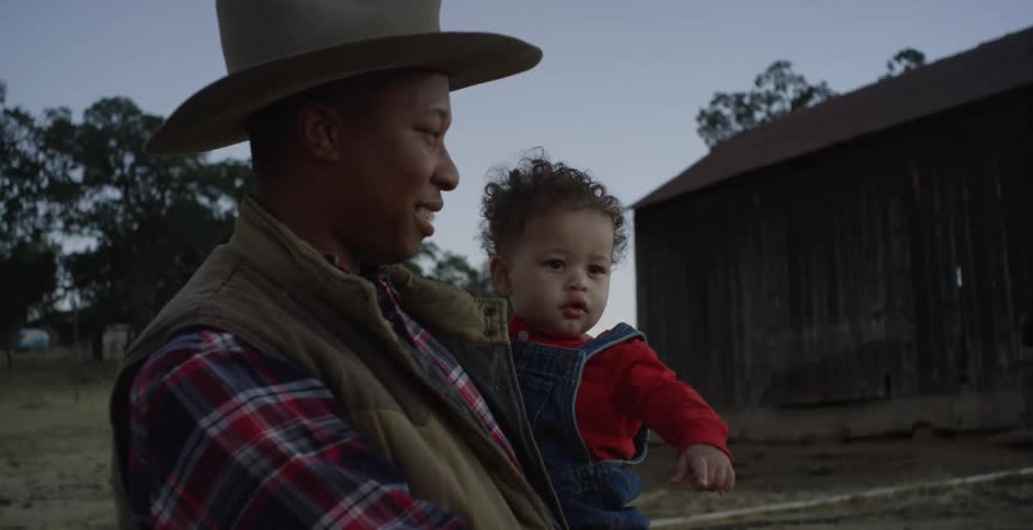 Mother wearing a cowboy hat holds her baby outdoors on the farm at evening