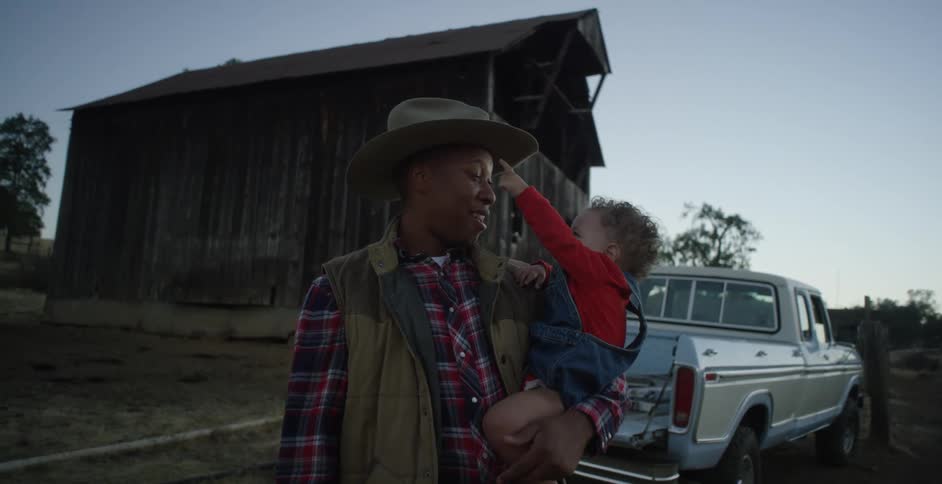 Father wearing cowboy hat and standing near pickup on farm smiles while holding child