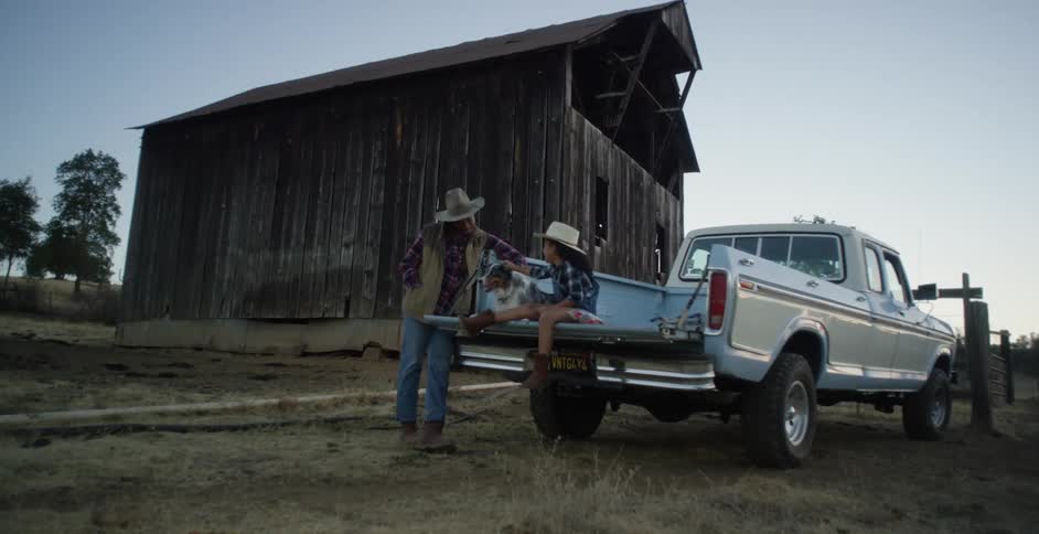 Father leaning against pickup truck on farm and daughter sitting in truck bed both interact with pet dog