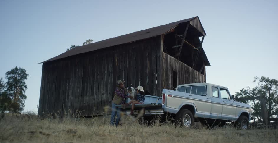 A man stands next to a truck that a little girl and dog are sitting on in front of a barn