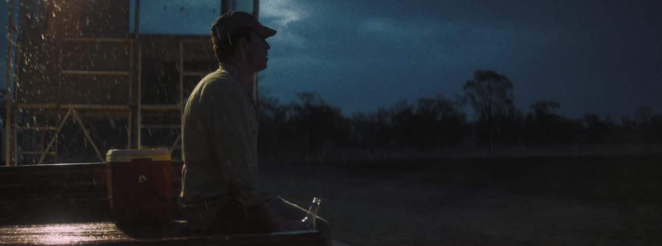 A man is sitting on the back of a pickup truck at night while it rains with a cooler next to him