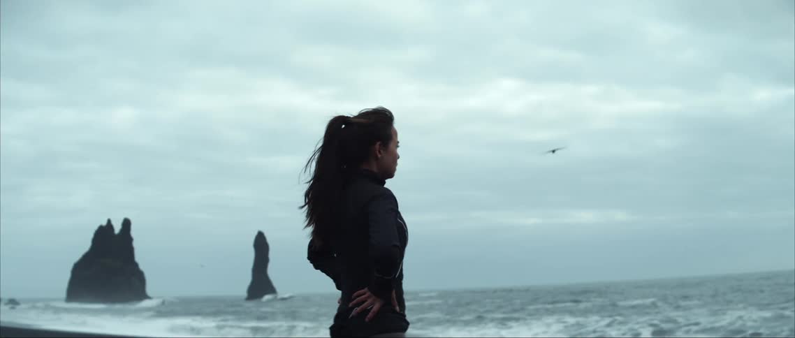 Woman stands on beach as waves roll past rock outcroppings under an overcast sky