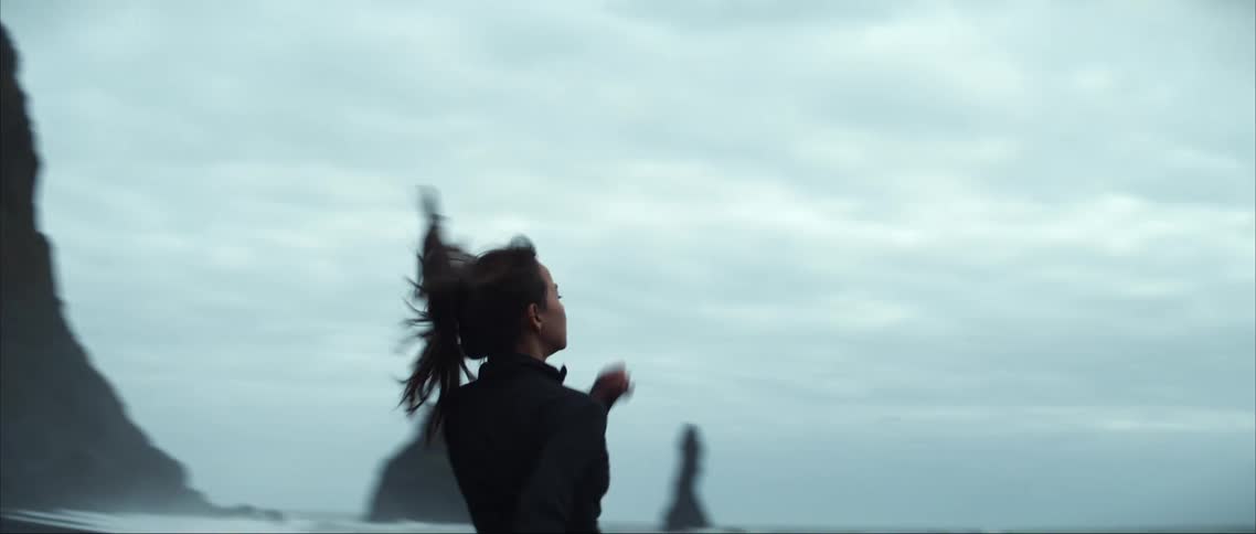 Woman running towards the rocky beach stops near the ocean and gazes in the distance on a cloudy day