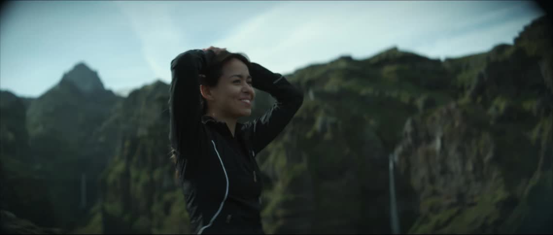 A woman runs to the edge of a cliff in a canyon and smiles wide at her accomplishment