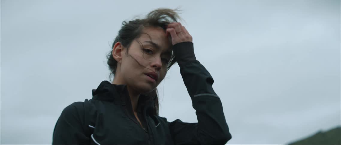 A woman is standing outside near a grass covered mountains as she moves her hair with her hand