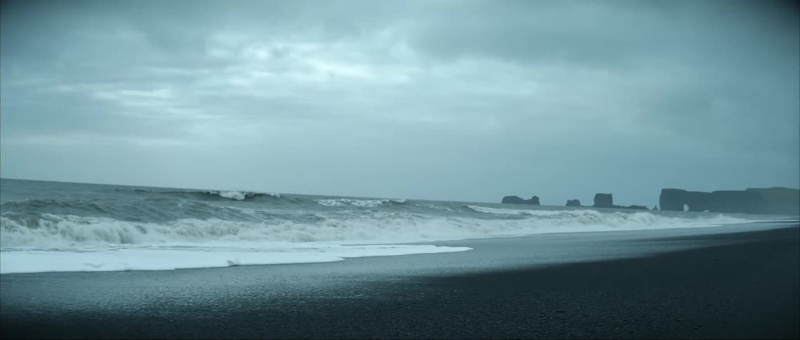 Rough water waves roll into shore and over the beach under a cloudy sky