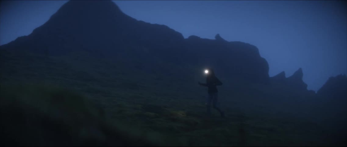 Woman wearing headlamp runs across foggy rough terrain near craggy mountains