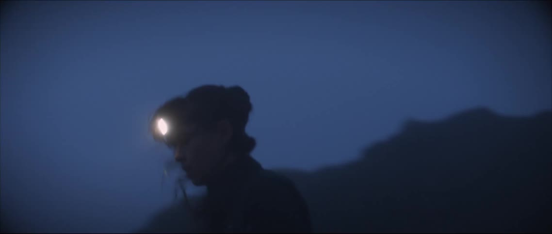Woman wearing headlamp moves across rough terrain at base of rocky hill