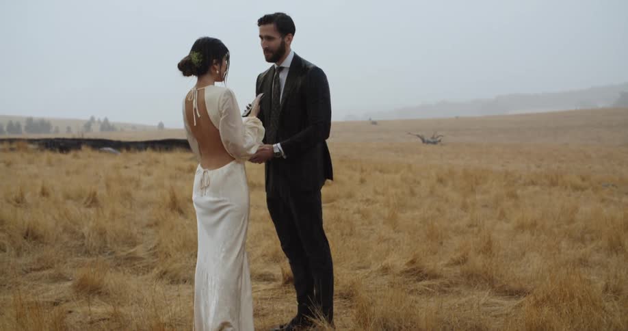 A man wearing a suit and woman wearing a long white dress are standing in the rain in a rural field as she holds her cell phone and he is holding her hand with both of his hands