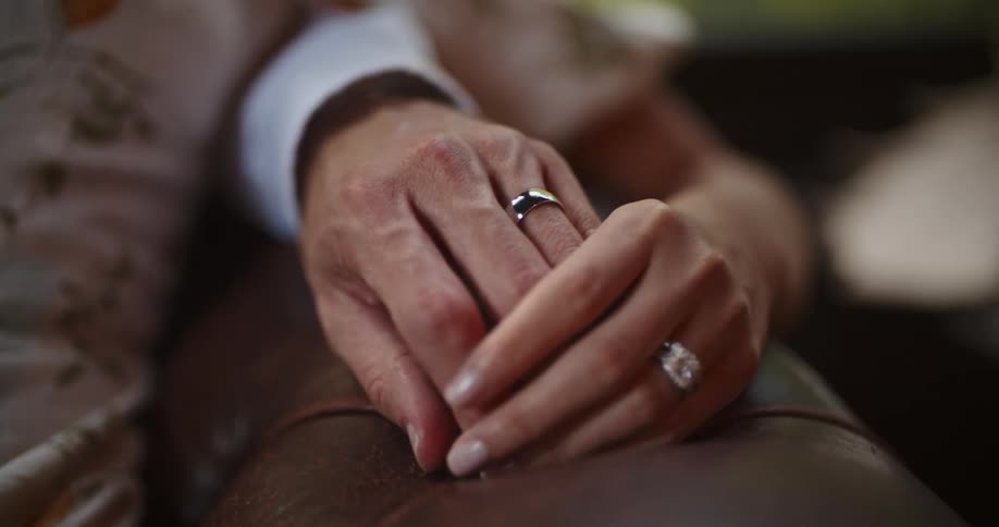 An elderly couple hold hands while sitting on the couch