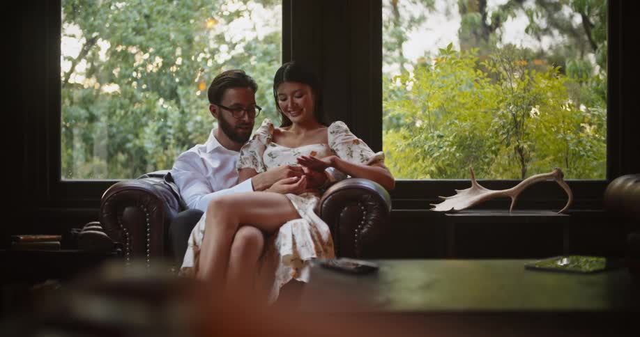 Woman with floral dress sits on mans lap sitting in brown armchair in the living room