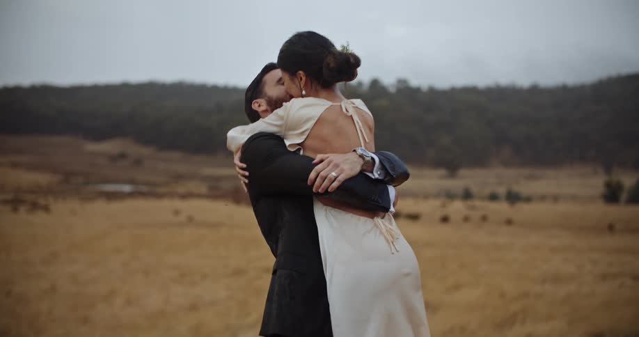 A bride and groom smile and hold each other while kissing outside
