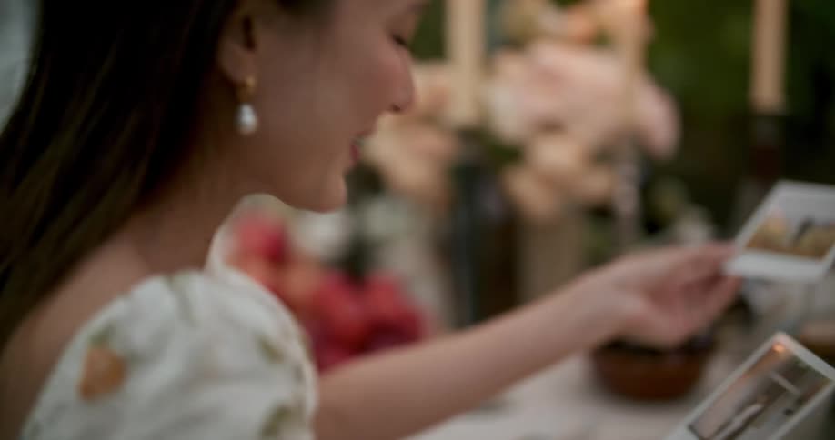 A woman is sitting at a table with candles holding a polaroid picture of herself and a man standing in a field and she hands it to the man in the picture