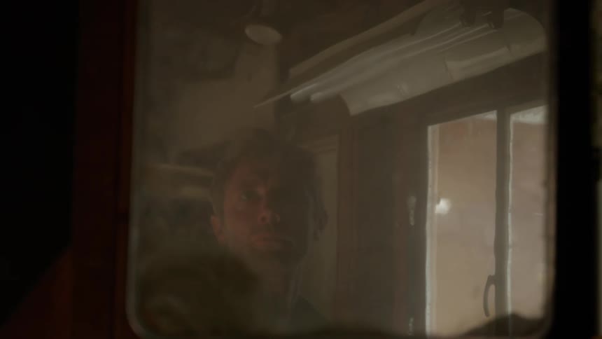 A man is standing in an old bathroom looking into the mirror and he opens the cabinet as he reaches in and holds a plastic bag