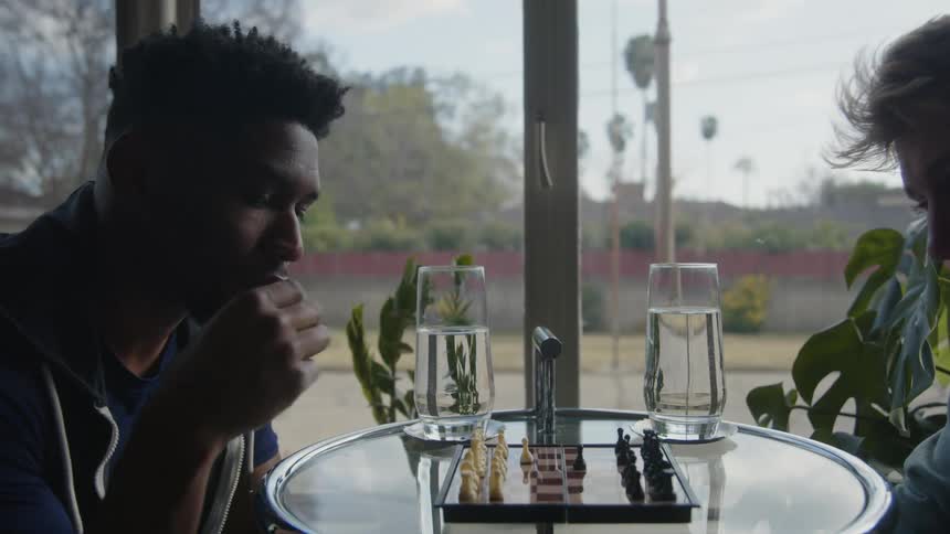 Two men are sitting at a glass table playing chess near a window with two glasses of water on the table
