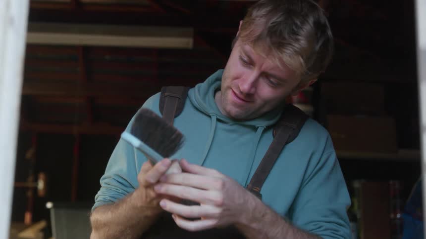 A man wearing a hoodie and overalls is holding a brush as he rolls up his sleeve and continues to work
