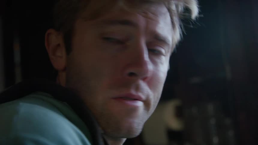 A man wearing a hoodie and overalls is standing near a window as he looks back and forth