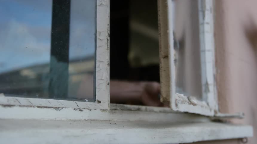 A man is standing inside a room in a building turning a handle to open the window