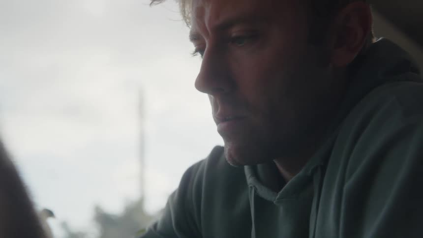 A man is sitting near a window looking ahead as he reaches his arm out