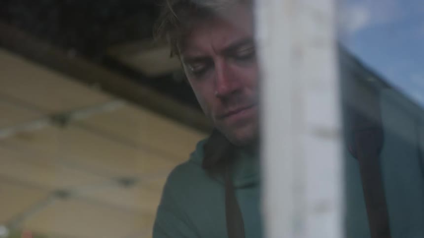 A man wearing a hoodie and overalls is standing near a window looking down as he works