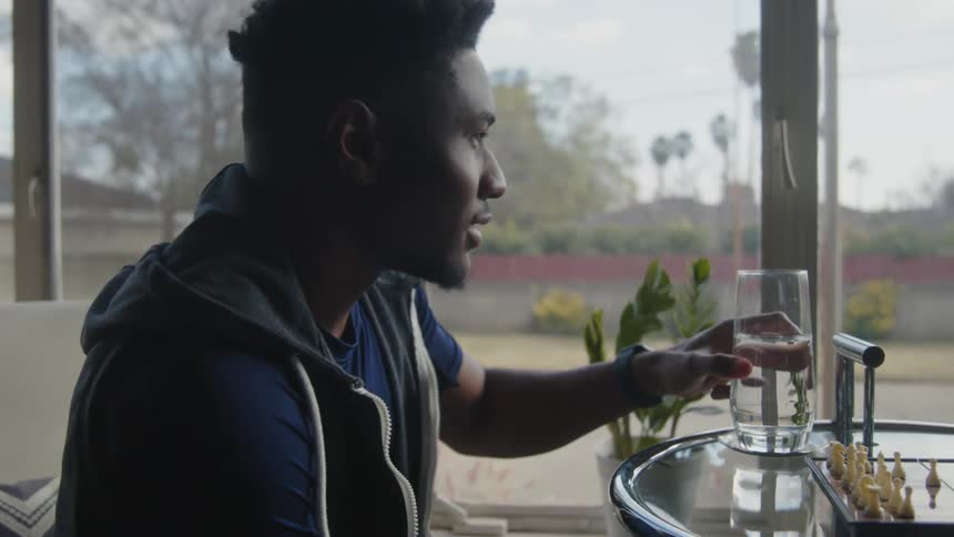 A man is sitting at a glass table playing chess near a window as he moves a piece and takes a drink of water the man on the other side is making a move