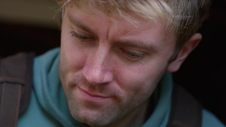 A man wearing a hoodie and overalls is standing near a window as he looks back and forth and smiles