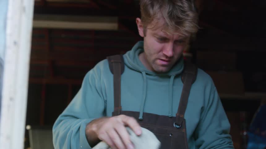 A man wearing a hoodie and overalls is standing near a window as he picks up a cloth and wipes the area in front of where he is standing