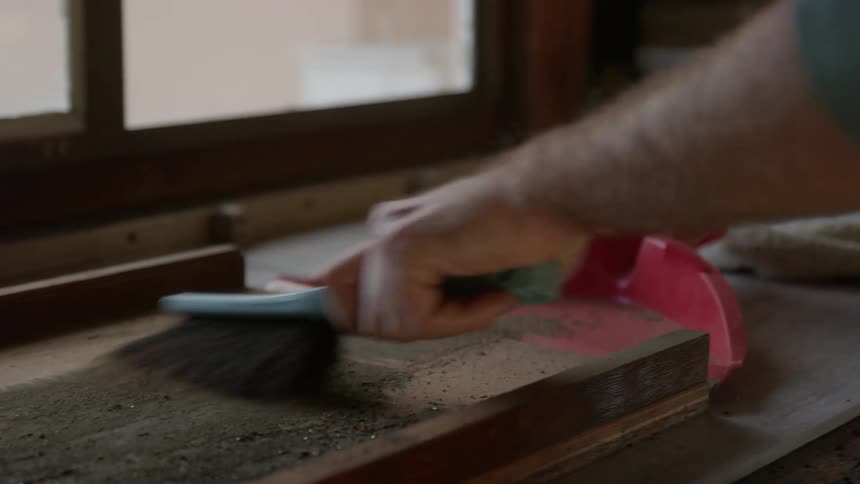 A man is standing at a table brushing dirt from a wooden board on to a dustpan