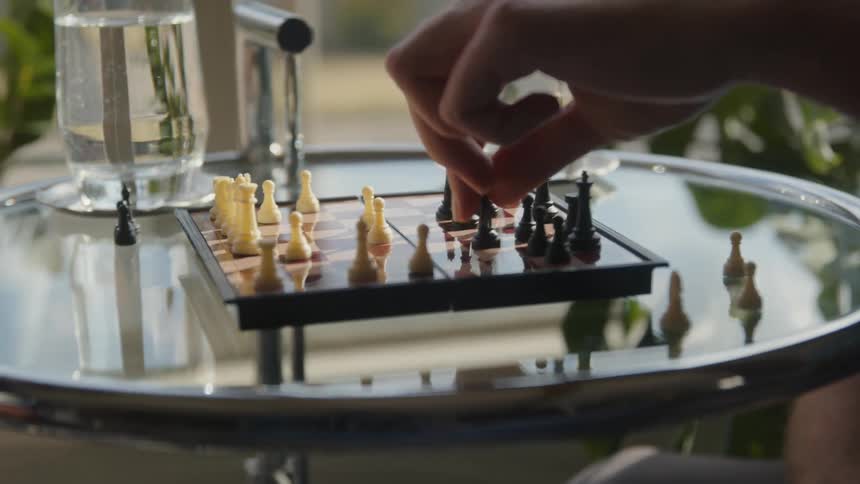 A man moves a chess piece over a small board over a glass table at home