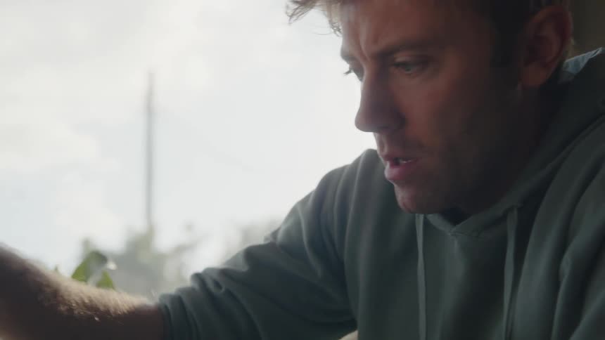 A man is sitting at a table playing chess near a window as he moves a game piece and laughs