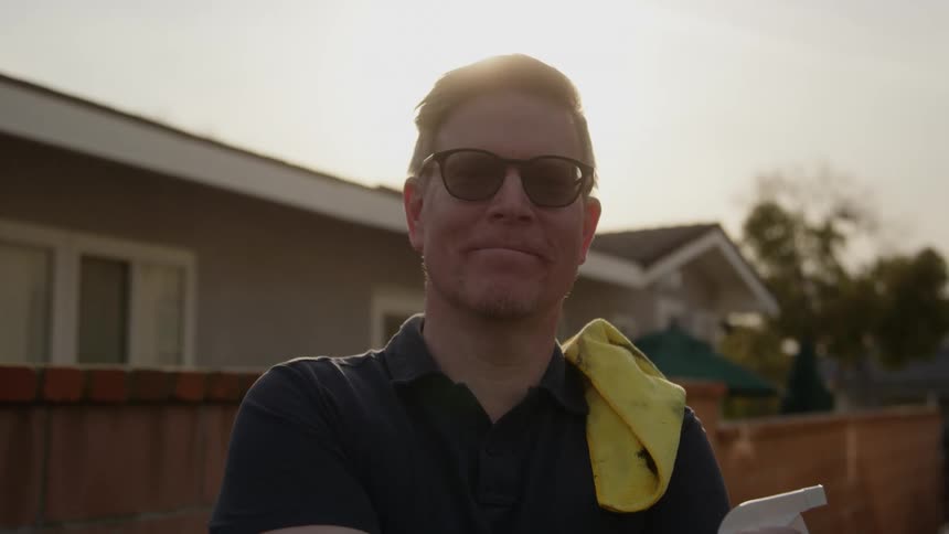 A man standing in front of a fence with a yellow rag on his shoulder smiling