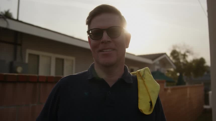 A man smiling outside a house with a towel on his shoulder on a sunny day