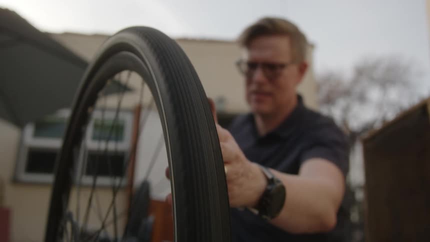 Man inspects bicycle tire outdoors with leafless tree in the distance