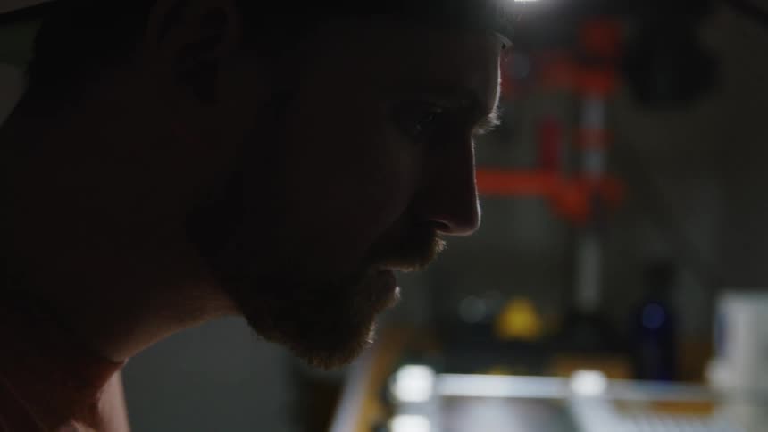 A bearded man leans forward over a workbench while concentrating on his task