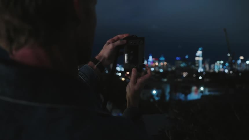A man is standing outside at night holding his camera and taking a picture if city buildings in the distance