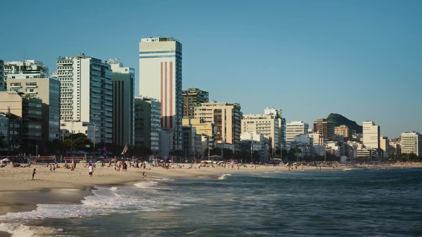 A large group of people are walking and sunbathing on a beach on a sunny day with hotels in the background
