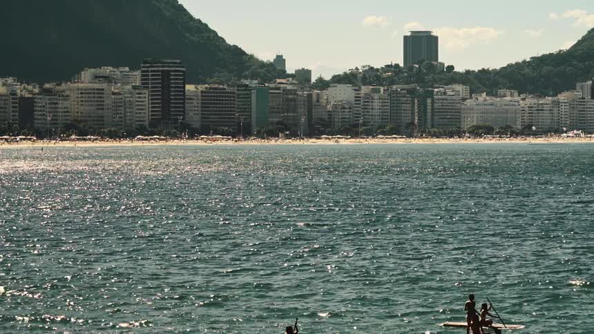 People ride paddle boards a distance from beach and city nestled at base of forest covered mountains
