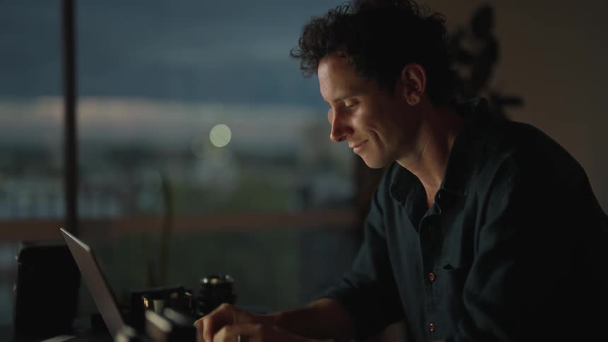 Man in black shirt types on laptop smiling in room at dusk