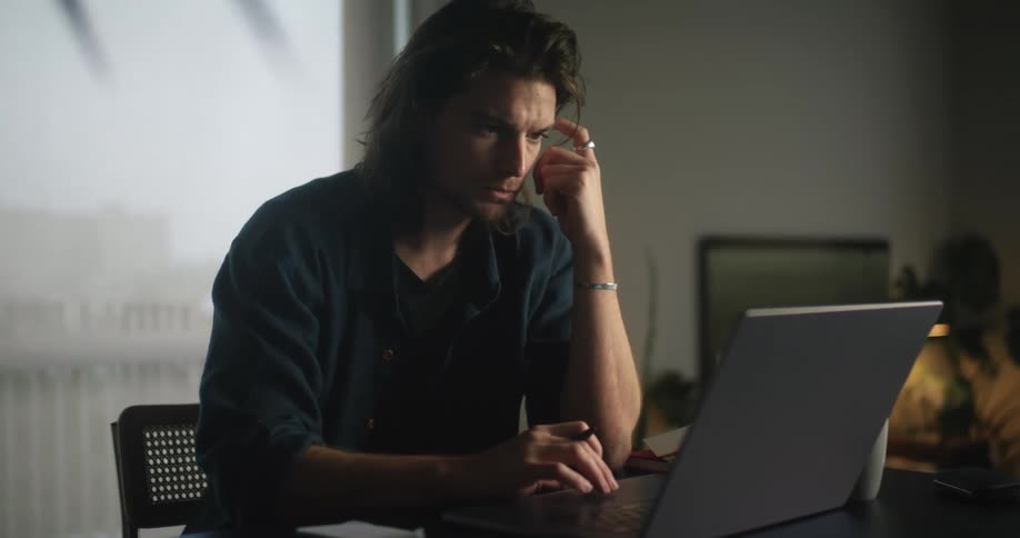 Man sitting at desk lookin at laptop reading emails and taking handwritten notes on small piece of paper