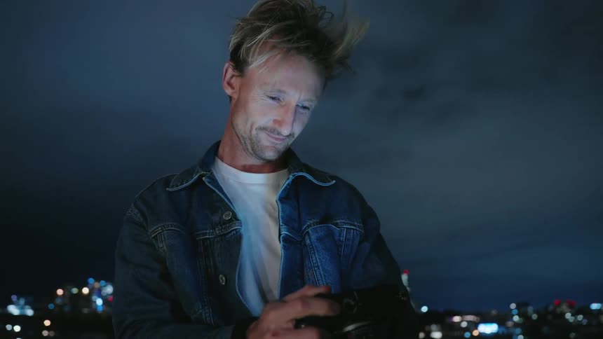 Blonde photographer checks his camera at night in front of the city skyline