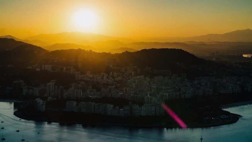 Buildings and towers stand tall in a city along the coast with boats sailing in the water and the sun shining brightly over mountains in the distance