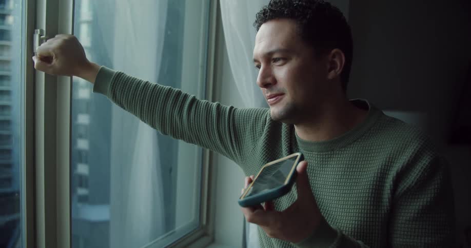 Man with green sweater talks on speakerphone looking out the windows at apartment building