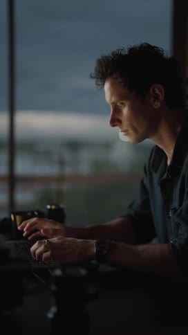 A man is sitting outside at a table looking at his camera lens and typing on a laptop computer