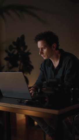 Man sitting at desk with laptop and dslr camera alternately typing on laptop and examining camera lens