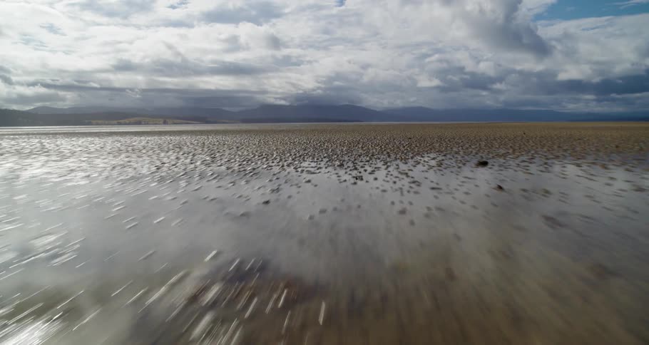 Moving along the shoreline in open rural land under a cloudy sky with mountains in the distance