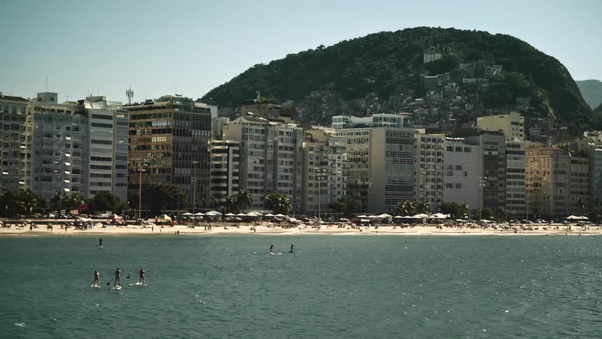 Buildings stand tall along the coast as traffic moves on the street and people swim in the water at the copacabana beach with buildings along the side of the mountain in the distance