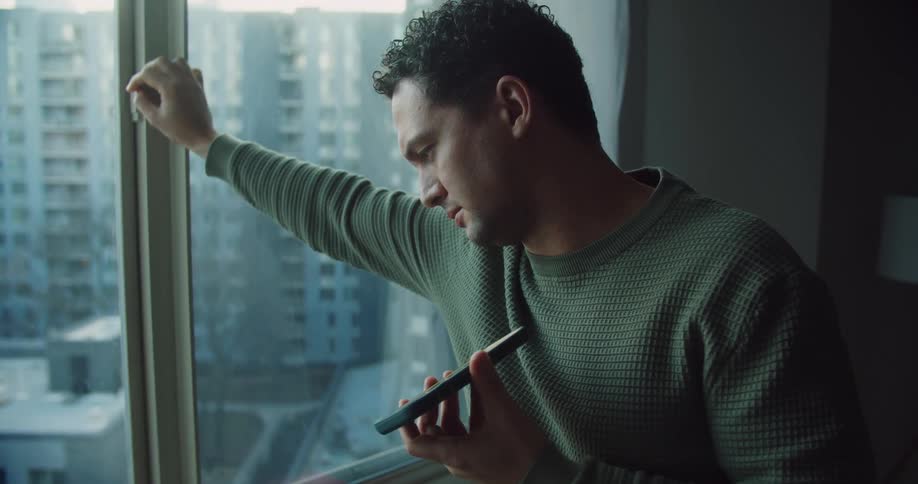 A man talks on the phone while standing next to his apartment window